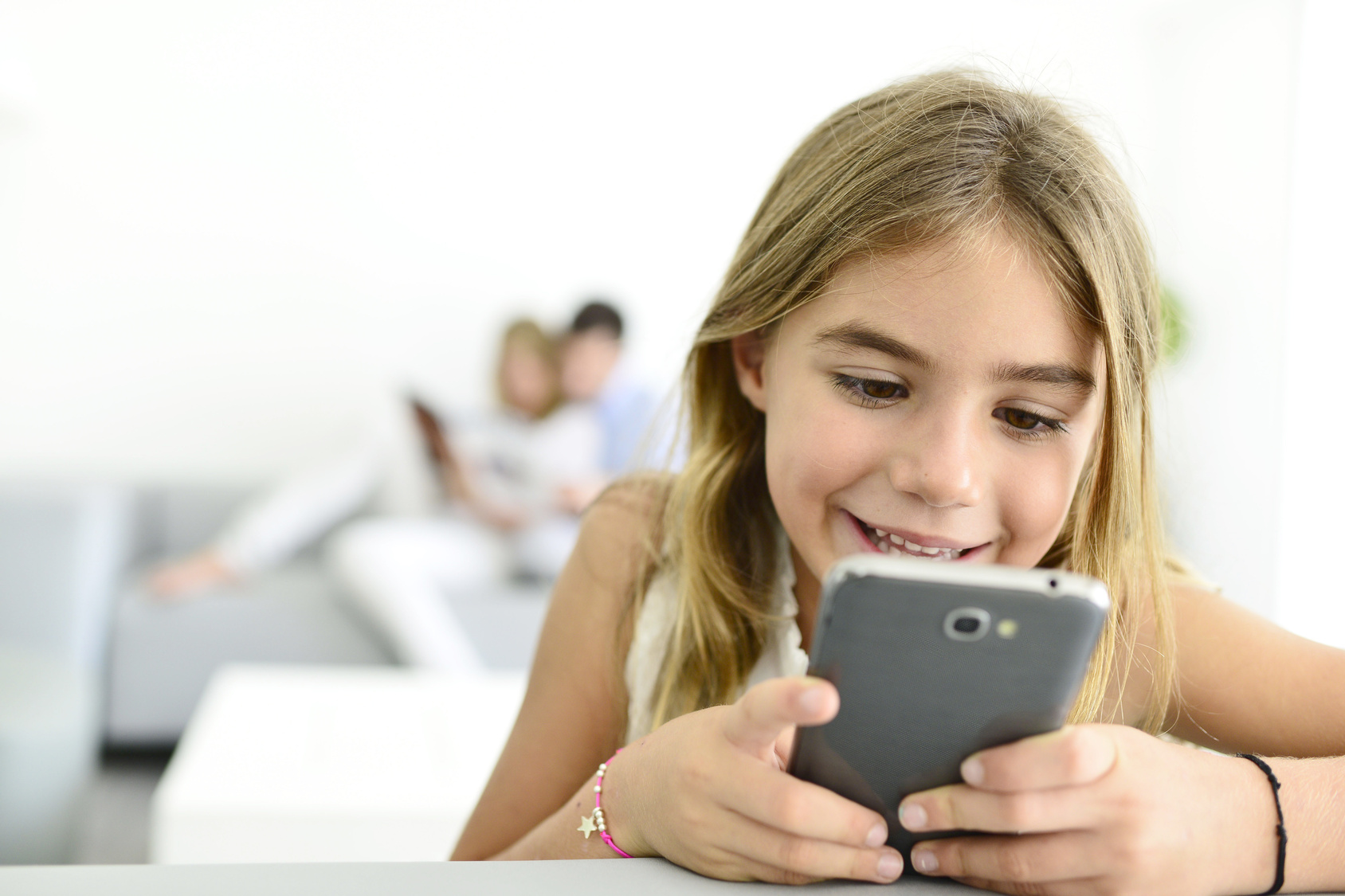  A smiling girl is holding a smartphone in her hands while her parents are reading a book in the background.